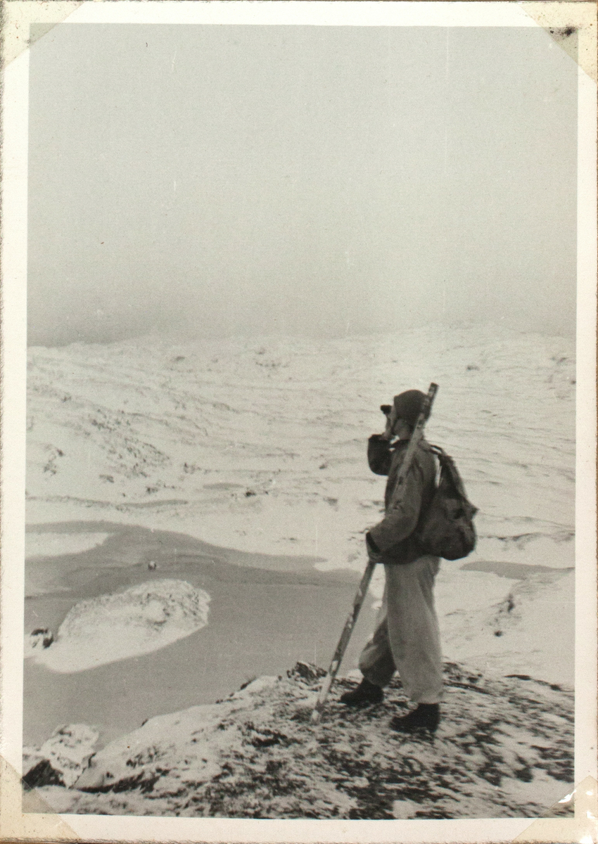 En Soldat står på snødekket Finnmarksvidda i det norsk-svenske grenseområdet og speider med en kikkert utover landskapet.