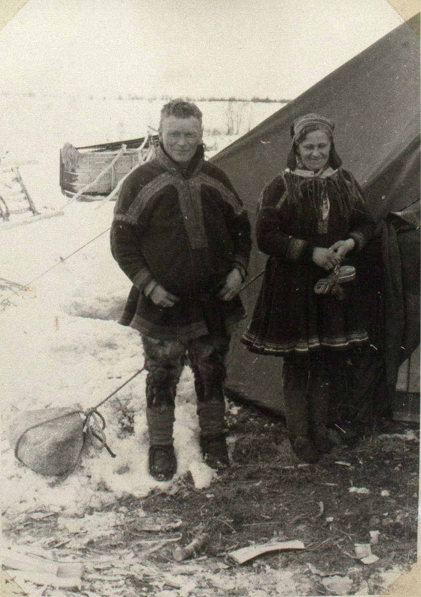 Bildet viser et ungt, samisk ektepar. De står ved siden av hverandre, foran et telt. De er ikledd tradisjonelle samiske antrekk. Teltet bak dem er festet med hjelp av stein. I bakgrunnen ser man en slede som ligger på side. Landskapet er snødekket.