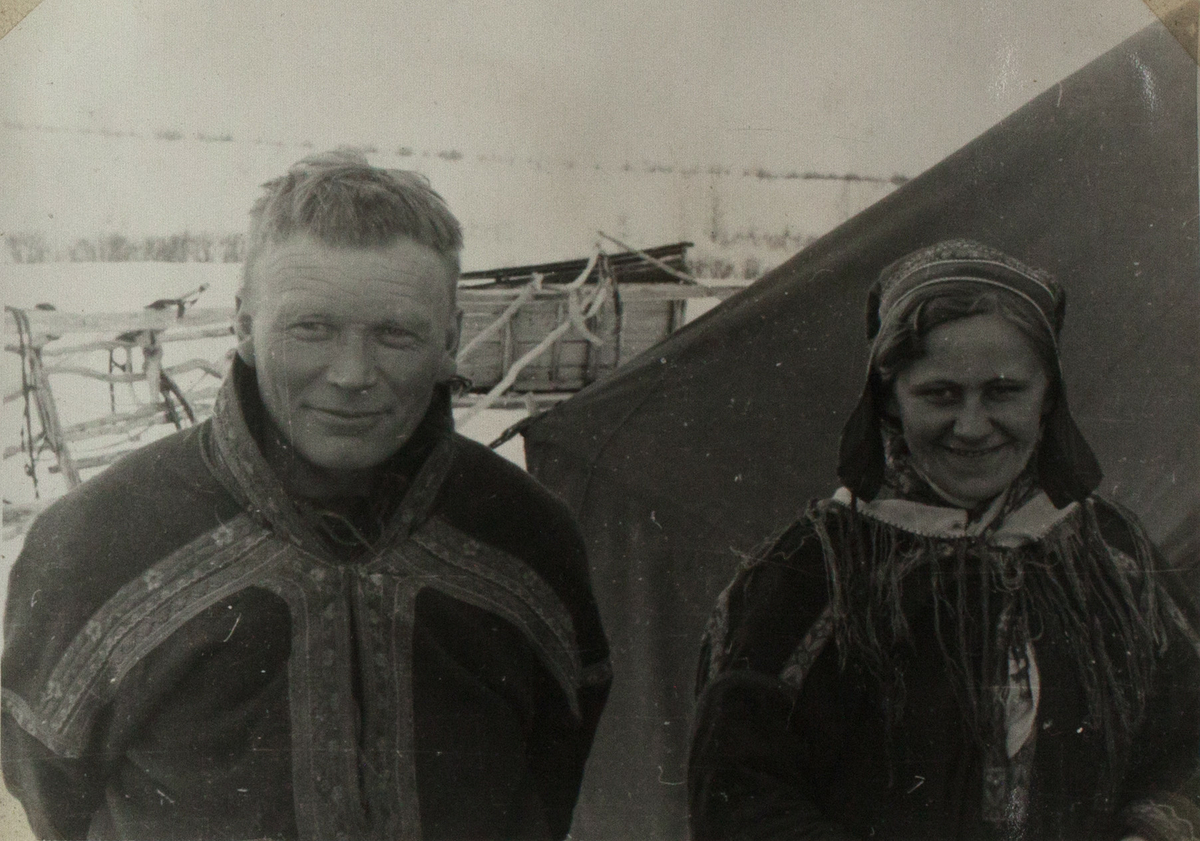 Bildet viser et ungt, samisk ektepar. De står ved siden av hverandre, foran et telt. De er ikledd tradisjonelle samiske antrekk. Begge kikker inn i kameraet og smiler. I bakgrunnen ser man en slede som ligger på side. Landskapet er snødekket.