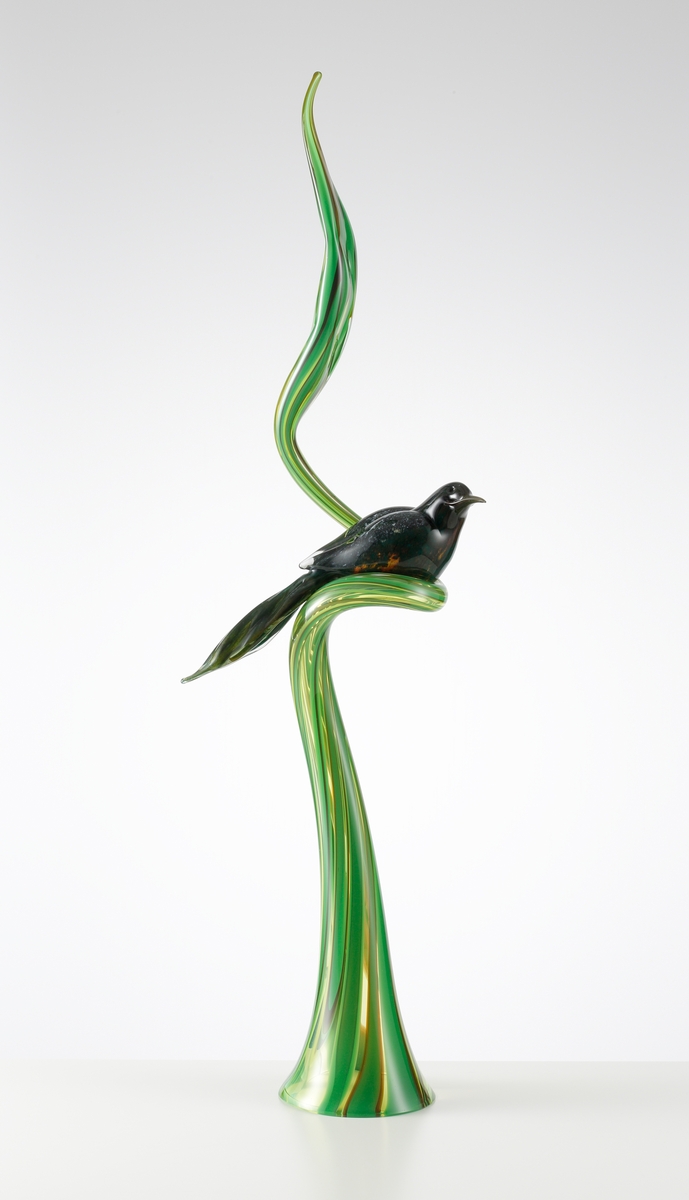 Skulptur bestående av ett en hög grön stående växt som avslutas med ett löv. Växten gör en sväng på mitten, varvid en exotisk, mörk och milerad fågel sitter.