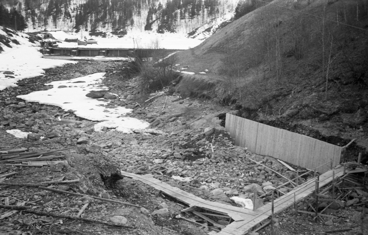 Anleggsarbeid ved Vinstravassdragets utløp av sjøen Olstappen i Skåbu i Nord-Fron kommune våren 1954, Her skulle det bygges en kraftig betongdam som ville gjøre Olstappen til et større vannmagasin for produksjon av elektrisk energi. Utløpet fra sjøen var stengt ved hjelp av en såkalt nåledam, som hadde vært brukt til å regulere vannføringa under fløting i det nedenforliggende vassdraget. I forgrunen ser vi treramper for trillebårtransport. Til høyre var det også reist en skjerm av tre, muligegns første byggetrinn i ei forskaling til en mur som skulle skjerme innløpet mot den nye dammen.