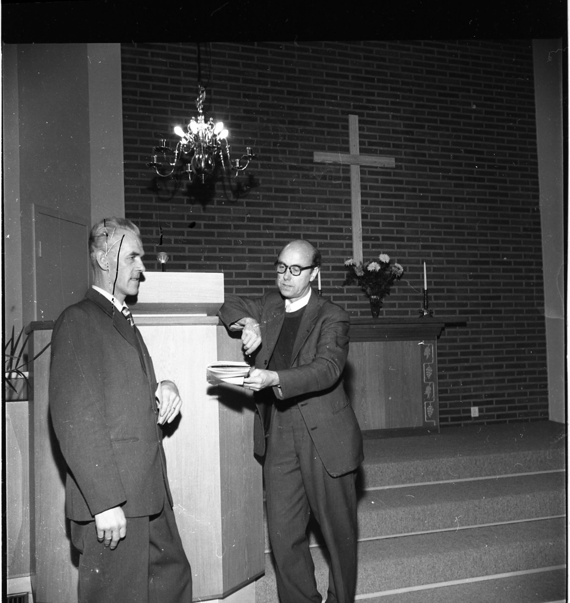 Två män står och hänger mot en talarstol längst fram i en kyrka. Den ena mannen håller fram en bok mot den andra. Bakom dem finns ett altare. På altaret står en bukett blommor.
