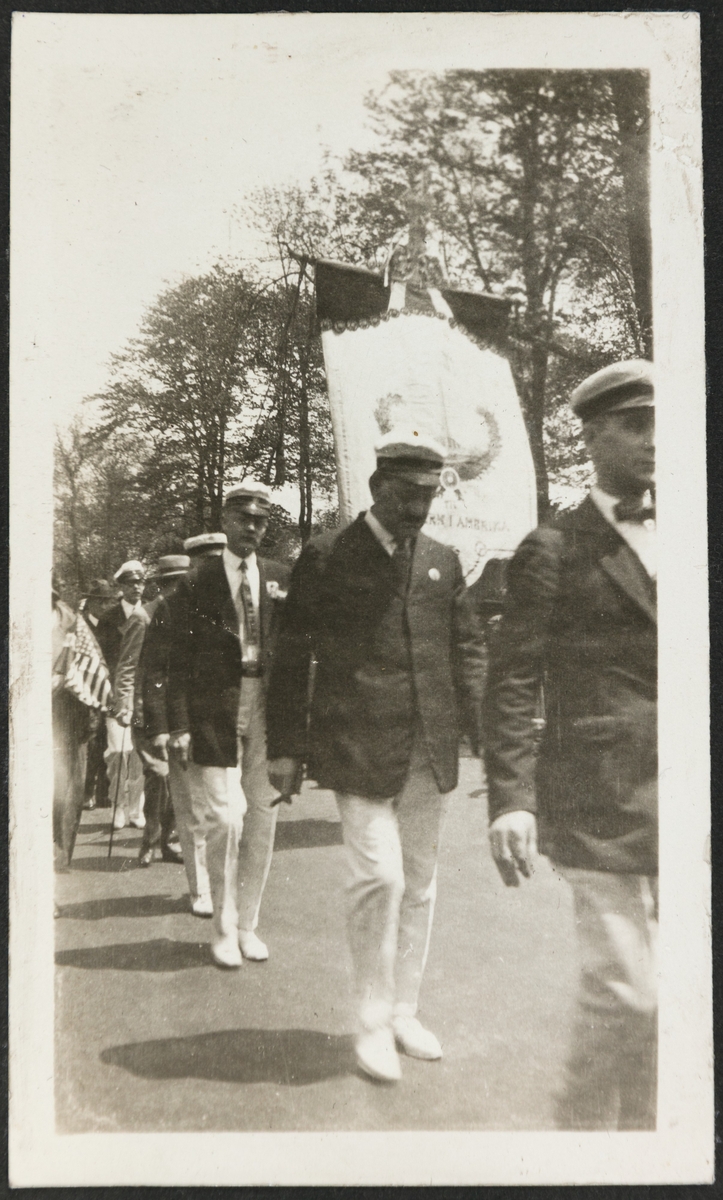 Syttende mai tog i USA. En gruppe menn i like klær går rundt ei fane med bilde av Haraldstøtta i Haugesund. Noe av teksten er synlig på fana: "TIL ...RK I AMERIKA".