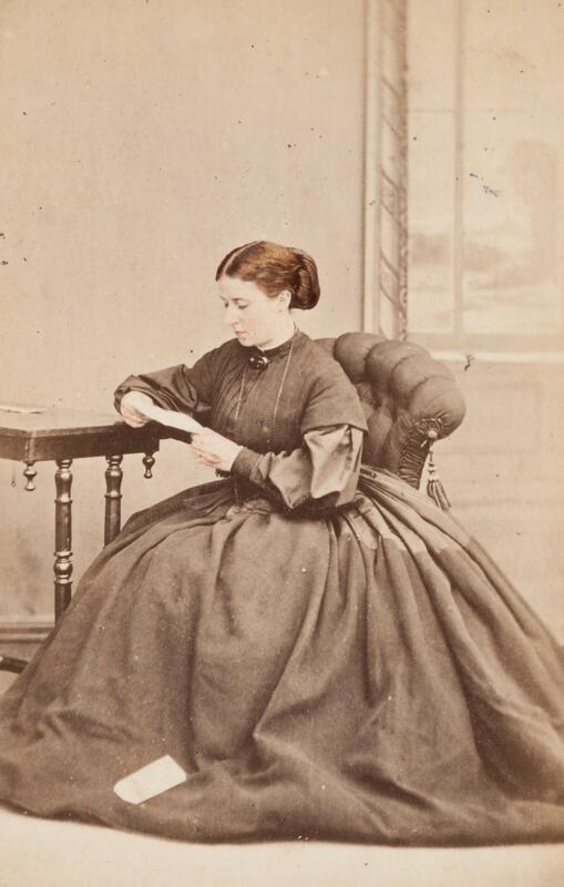 En kvinne i en stor kjole sitter ved et bord og leser et brev.