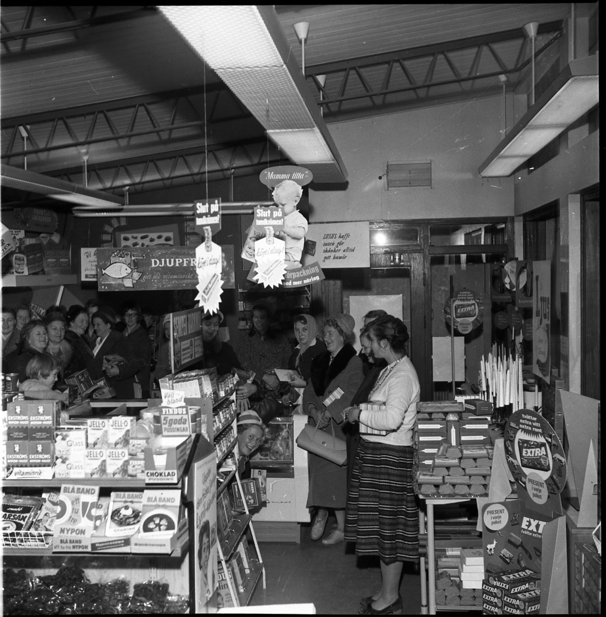 Kvinnor står i en vid båge i en mataffär. En kvinna tittar fram bakom en hylla och ler mot kameran. På hyllorna står matvaror med varumärken som Ekströms, Blå band och Jell-o.