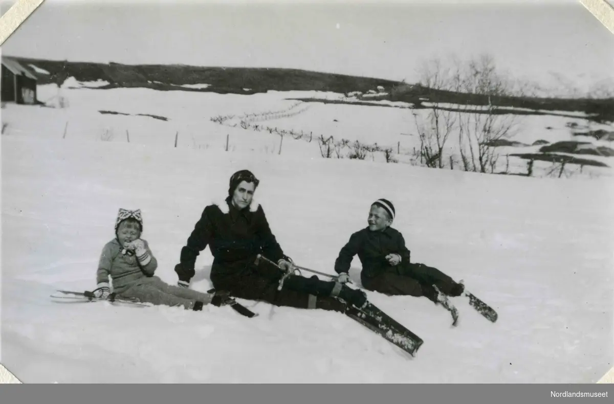 En kvinne og to barn sitter i et snødekt landskap med ski på beina. Barna har strikket lue og votter.