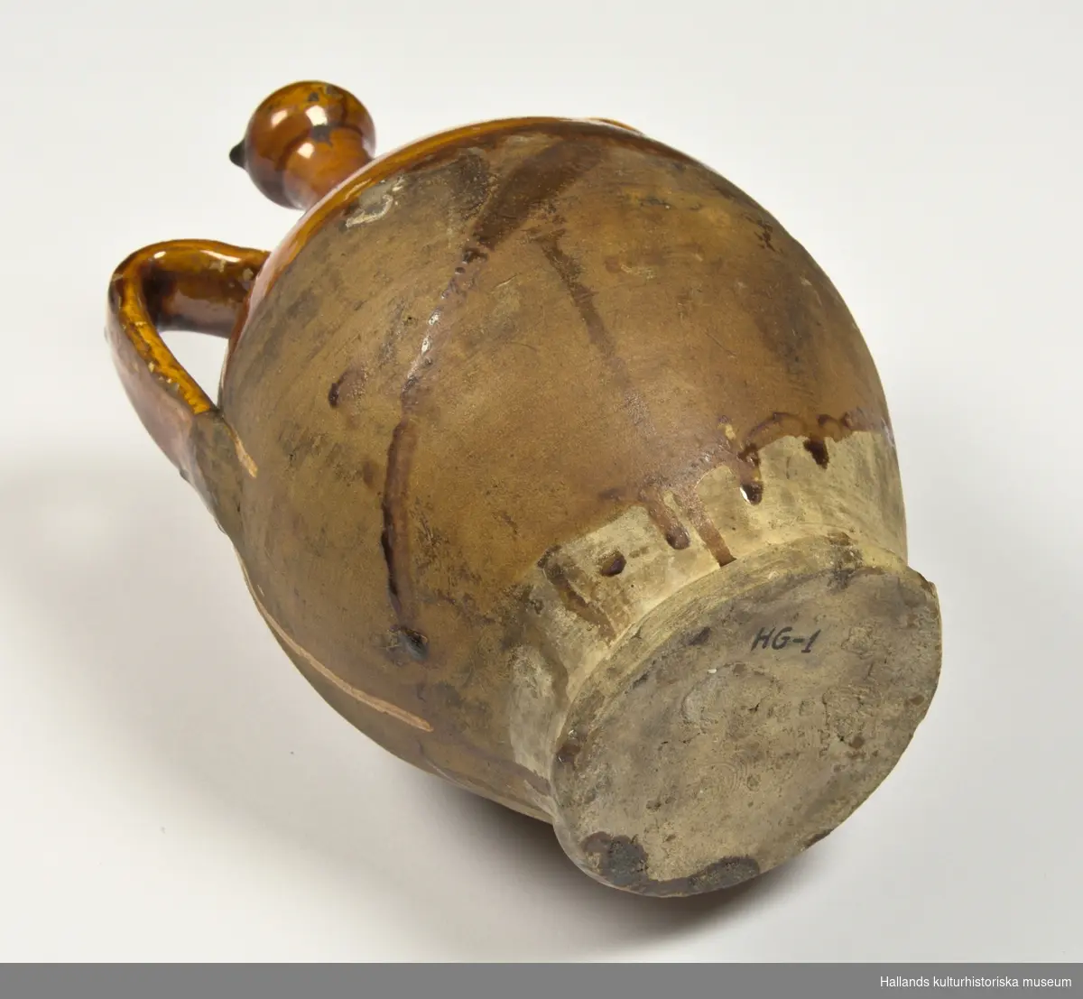 Krus, (urna) av keramik. Har två öron, (handtag) och smal hals. Med glaserad överdel, gulbrun, flytande glasyr.