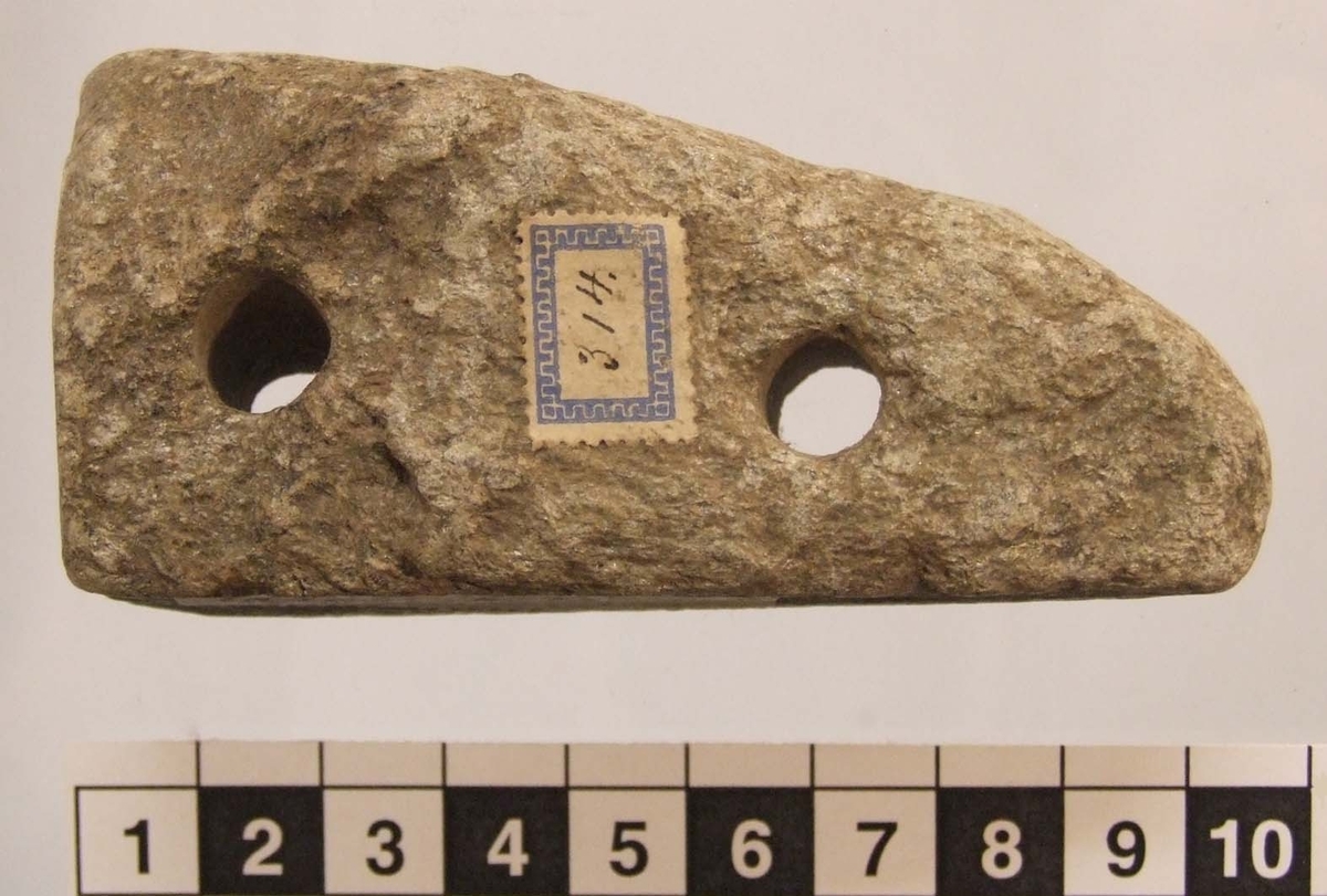 Sten med två plana sidor och två hål borrade genom stenen, 11 mm diam. Sannolikt ej förhistorisk.