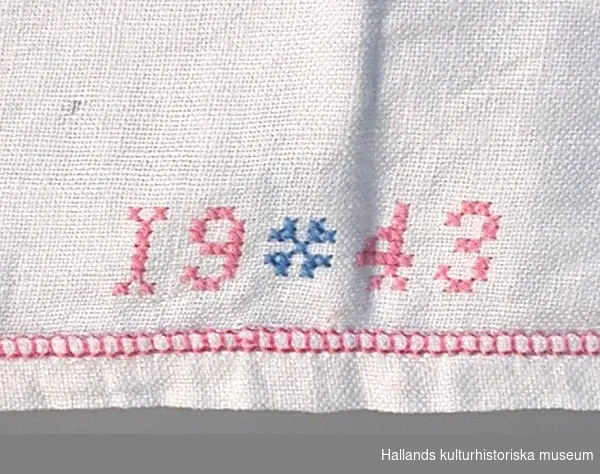 Duk av tuskaftat linne, broderad med korsstygn i rosa och blått samt utdragssöm sydd med rosa garn. Märkt: "AA 1943".