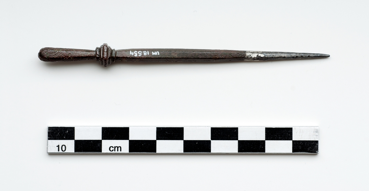 Föremål i metall, kopparlegering. Stylus med sexkantigt tvärsnitt. Spetsen av vitmetall, eventuellt silver. Motsatta änden profilerad samt skedformad. Möjligen örslev.