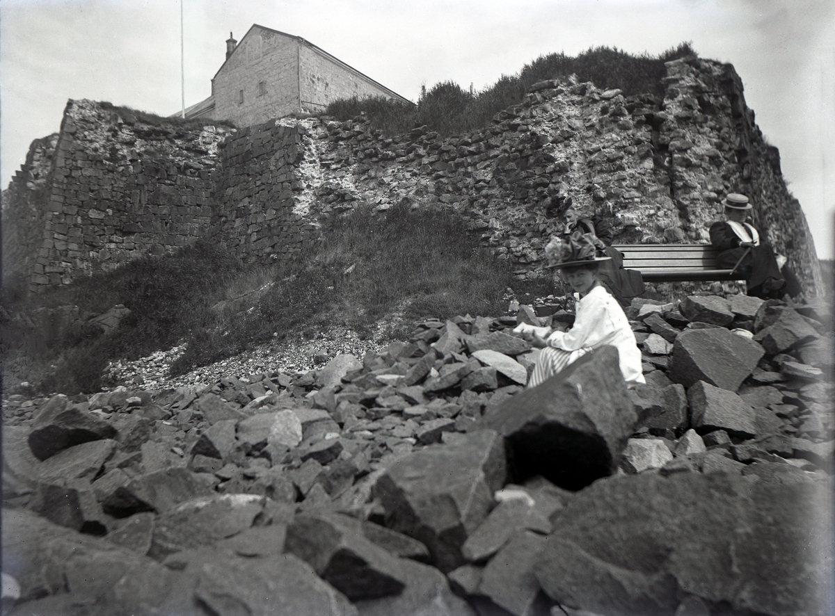 Foto av tre kvinnor som slagit sig ner framför fästningsmurarna till Varbergs fästning. En yngre kvinna sitter bland stenskravel, de andra har slagit sig ner på en parksoffa.