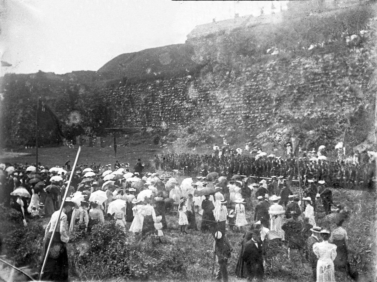 Mängder av folk har samlats på gräsplanen nedanför Varbergs fästning.
Närmast fästningsmurarna ser man bl a en pluton beväringar.