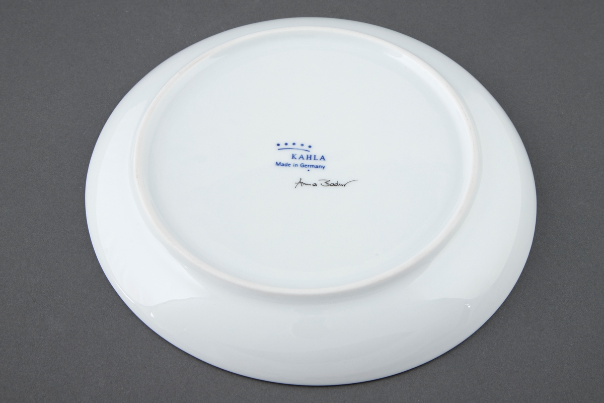 Rund tallerken i hvit porselen med utflytende blå underglasur i bunnen. Svak helling på fatet slik at det er høyere på den ene siden.