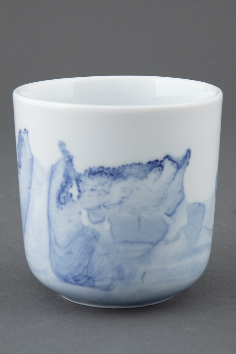 Hvit kopp med rette vegger. To tredjedeler av koppen er dekt av et transparent bølgemønster i blå underglasur.