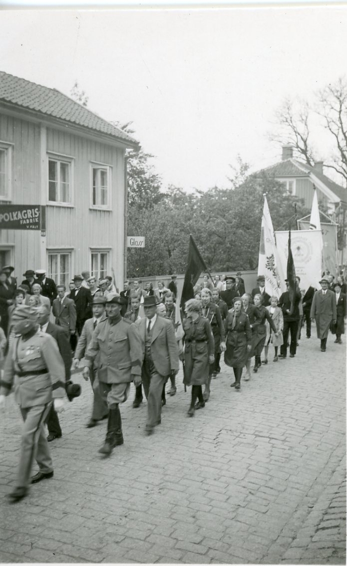En kortege med militär i täten tågar Brahegatan söderut i Gränna. En fanborg följer efter. En fastighet till vänster skyltar med "Polkagrisaffär V. Fält"