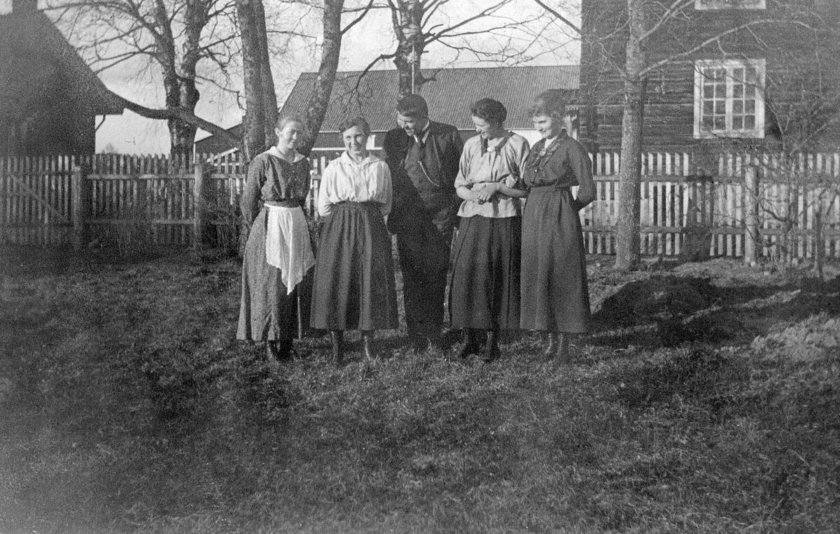 Gammelt foto av fem personer som står ved siden av hverandre i en hage. En mann i midten og to kvinner på hver side.