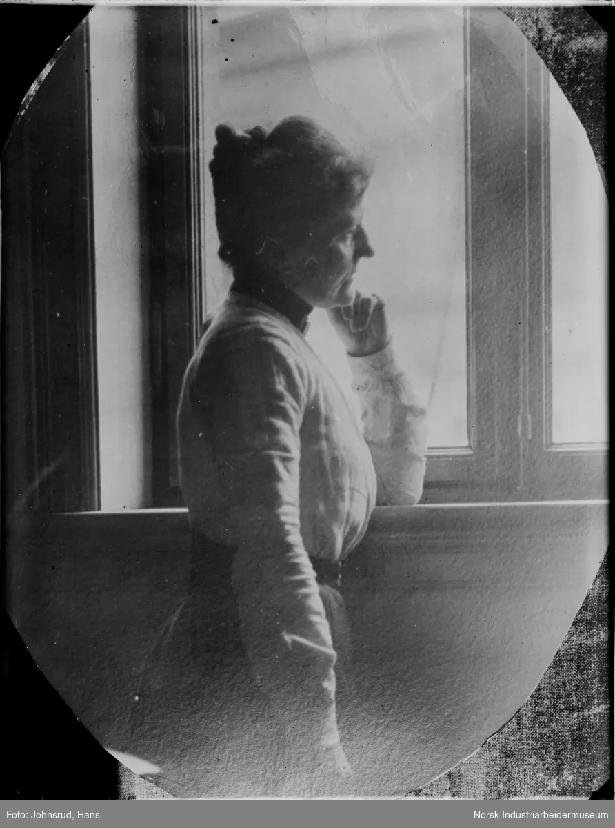 Portrett. Kvinne stående foran vindu, hun lener seg med albuen i vinduskarmen.