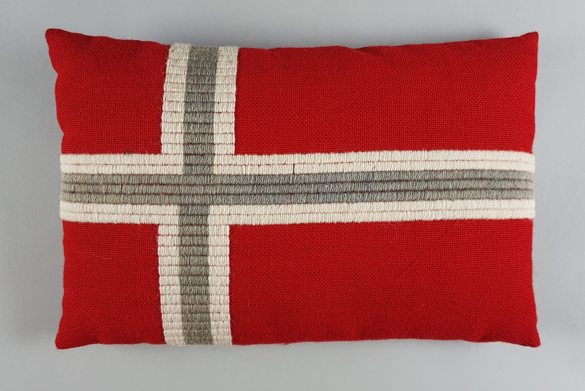 Brodert pute med norsk flagg, i rødt, hvitt og grått (falmet blått?). Grovt vevet ullstoff med hvit og blå broderte striper.