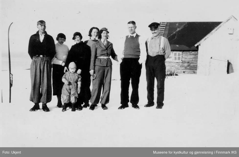 Bildet viser en gruppe av åtte personer som står i snøen foran et hus. Det er fint vær. De er et barn, fire kvinner og tre menn. Navnene dere er ukjente. Til venstre stikker det en ski ut av snøen. Bildet er trolig tatt på Melkøya i Finnmark i 1936. 
