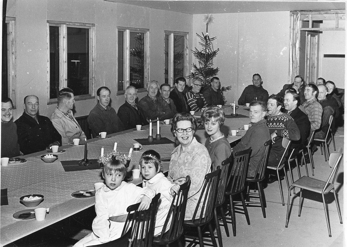 Ett stor antal män, två kvinnor samt en liten lucia och tärna sitter runt ett kaffebord med tända ljus. I bortre hörnet står en julgran.