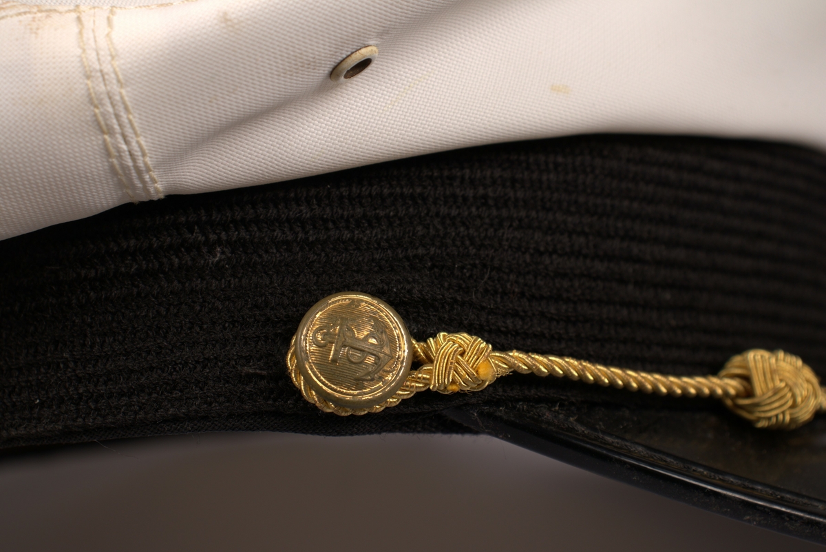 Uniformslue med hvit pull og svart skjerm. Luemerke med rederiflagget til Ofotens Dampskibsselskap. Gull snor festet over skjermen med to gullknapper på hver side.