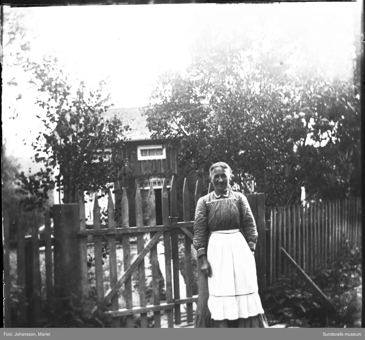 En kvinna med vitt förkläde står invid grinden till ett bostadshus. På bild nummer två har hon en katt i famnen.