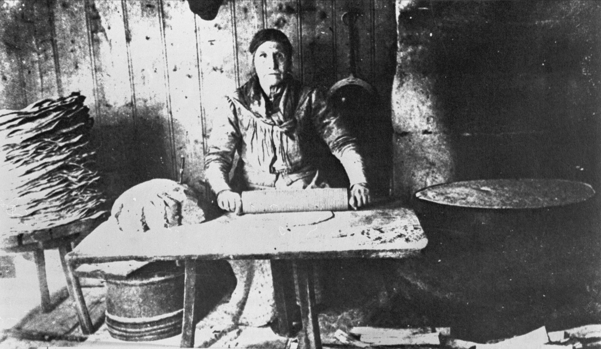 Eldre kvinne ved bakstebord. Til venstre en stabel med flatbrød, til høyre en steketakke.
