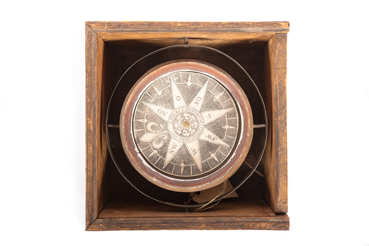I kvadratisk låda av al, omålad, utan lock. Kardanupphängd kompass. Kompassskål av svarvat trä, omålad, invändigt vit med röd överkant. Streckindelad kompassros.