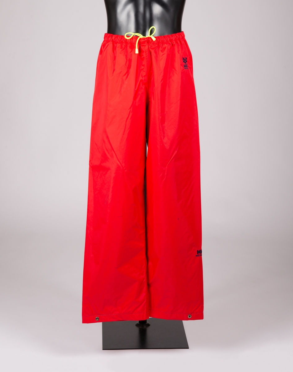 Art. nr. G-474. Rød bukse med mørkeblått fôr med HH Helly Hansen logo som mønster. Gult snortrekk med dobber i livet, i tillegg til elastikk. Knapperegulering med to knapper nederst på beina. Hempe innvendig bak med ved HH-logo.
