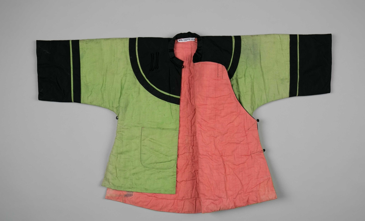 Kinesisk vattert omslagsjakke for barn (4-5 år) i grønn silkebrokade, kantet med svart brokadestoff. Lys rosa silkestoff i linningen (opprinnelig rød?). Lukkes med tre hemper. En utvendig lomme.