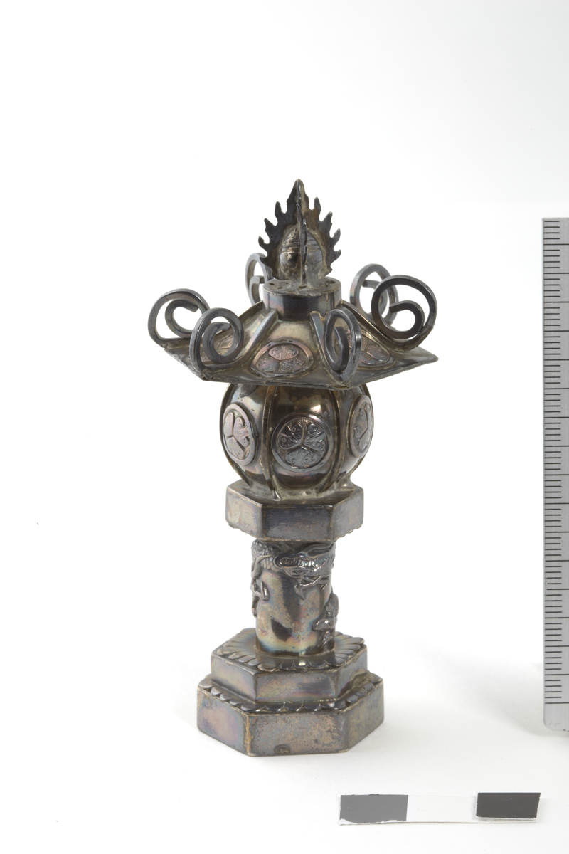 tempellampe. lampeholderen er rund med 6 felter, i hvert felt et tokugawa-våpenmerke, over denne en sekskantet hatt med 6 tokugawamerker og seks spiraler. en drage