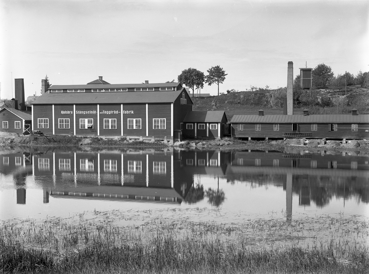 Vy mot Hjulsbro Tråddrageri- och Spikfabriks AB. Verksamheten startade 1907 invid fallet i Stångån vid sidan av Hjulsbro sluss. Efter konkurs 1921 uppköptes företaget av den driftige entreprenören Jonn O. Nilson, som utökade produktionen med stängselnät och taggtråd i särskild fabriksbyggnad. Här en vy mot den nya anläggningen något av 1920-talets senare år.