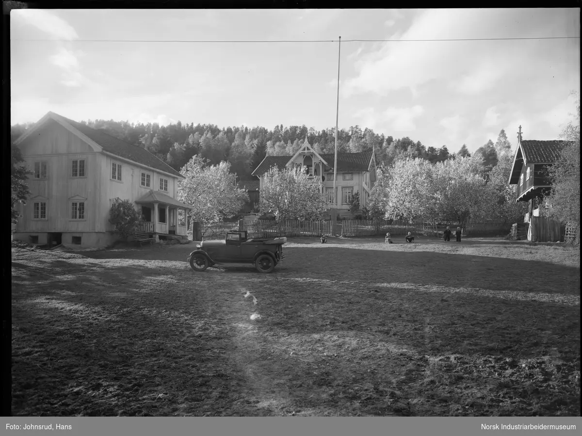 Gårdsplass på gården Seljord i Heddal med to bolighus og ett stabbur. Barn sittende på gresset på gårdsplassen. Bil stående foran ett av bolighusene. Flaggstang midt på tunet. Ett av bolighusene er dekorert med snekkerglede.