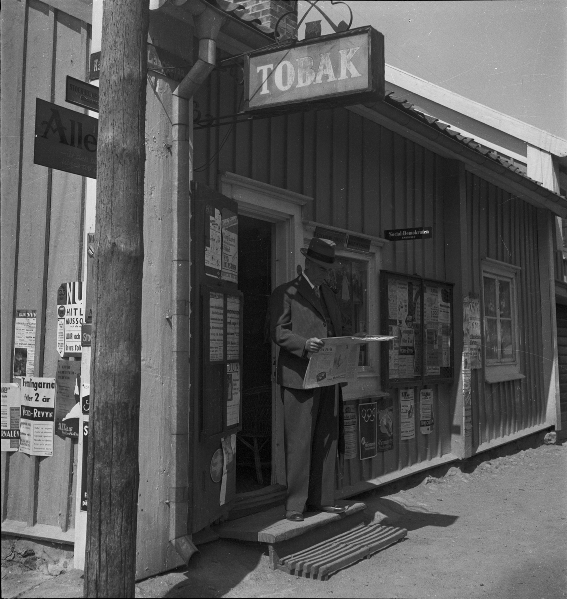 Man står på trappan till tobaksaffär och läser en tidning, Långgatan, Mariefred, 1936
