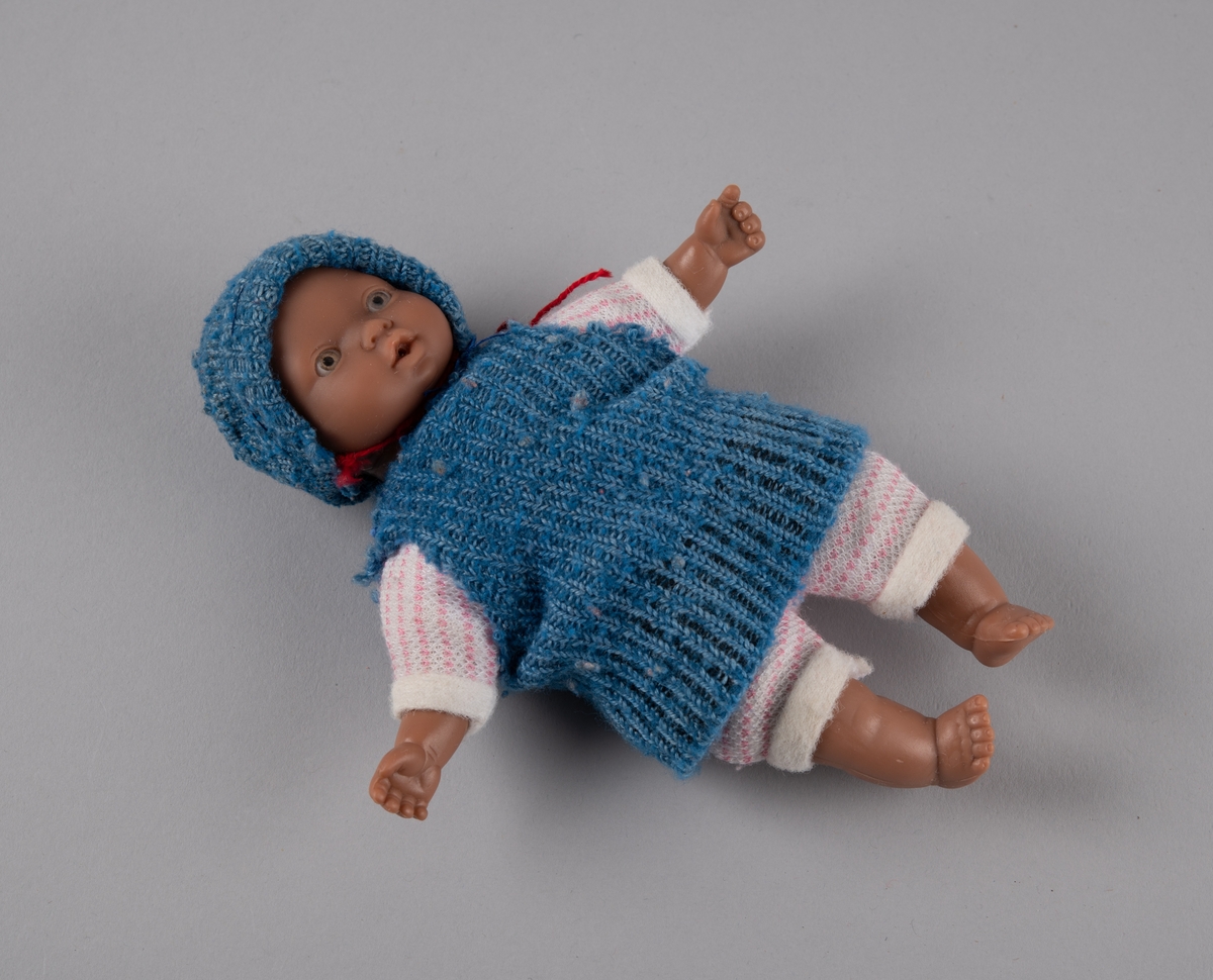 Babydukke med hode, armer og føtter av plast, og torso av tekstil fylt med vatt. Den er kledd i hjemmestrikket, rosa og hvit buksedrakt, samt blå genser og lue. Lua har en rød hakesnor.