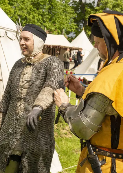 Ridder i platerustning og gul kjortel hjelper en annen ridder i ringbrynje med å feste ermene