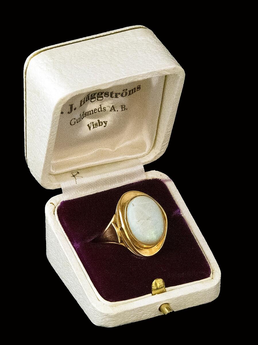 Ring, fingerring, av guld, med en oval slipad halvädelsten i ett upphöjt genombrutet fäste. Stenen gråvit med grön glans, sprucken, har lagats. Stämplar CJH (Carl Johan Häggström). Kattfot,18K, W, D10 (1978). Förvaras i ask av papp klädd i vit skinnimitation. Knäpplås. Invändigt lila sammet och trycket "C.J. Häggström Guldsmeds AB Visby".
I samlingen utan nummer