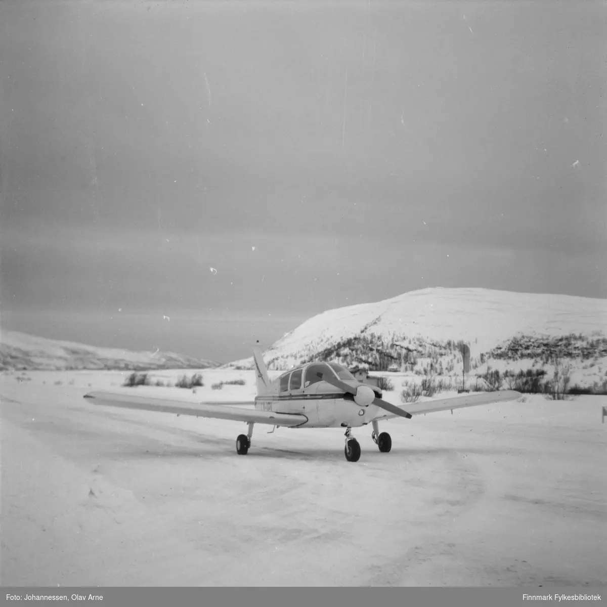 Ivar Bryggari i sitt Piper Cub fly på den private flyplassen i Båtsfjorddalen.

Flyet kan være modell Piper PA-28-180 Cherokee D. Flyet har ID nummer LN-HHH skrevet på venstre side (synes ikke på foto)

Flyet er omringet av sne og man ser vinterlandskap i bakgrunnen 