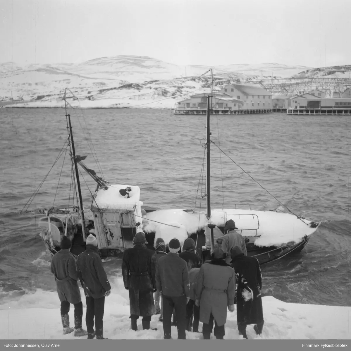 Foto av en gruppe mannfolk i vinterklær som ser på et havarert fiskeskip  

Sjark i land, på andre siden, på storholmen, Fuglevikbruket til venstre bak, så Aarnesbruket i Båtsfjord 
