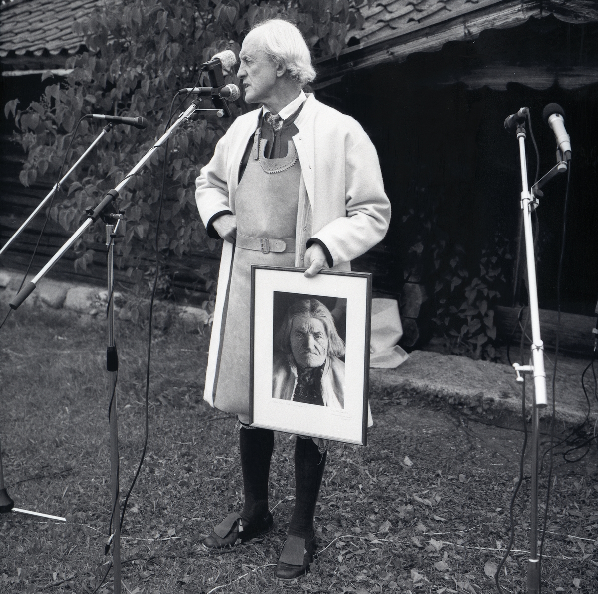 En man i folkdräkt står vid en mikrofon och håller en tavla föreställande ett kvinnoansikte under Lärkadagen på Sollerön den 22 juli 1990.
