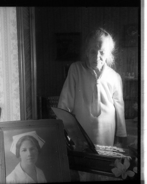 En äldre kvinna, till hälften i skugga, syns i en spegel ovanför ett fotografi på henne själv i unga år som sjuksköterska.