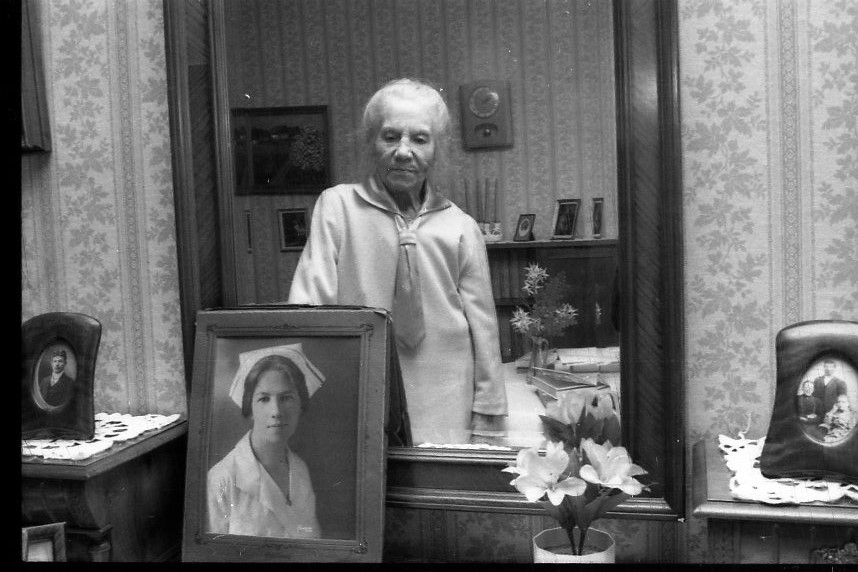 En äldre kvinna syns i en spegel ovanför ett fotografi på henne själv i unga år som sjuksköterska.