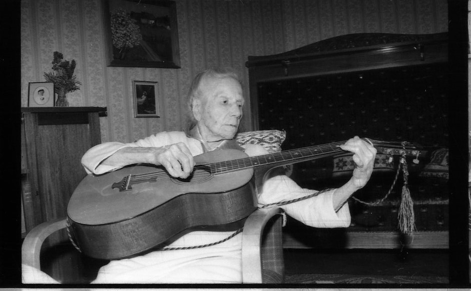 En äldre kvinna sitter tillbakalutad i en fåtölj och spelar gitarr.