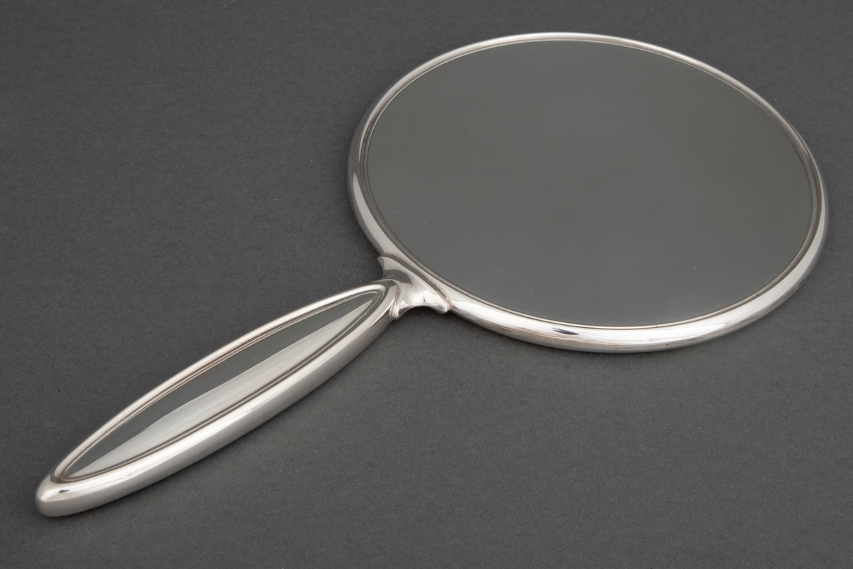 Sirkelrund speil med håndtak. Speilet er innfattet i sølv og har flat bakside med en profilert rand. Håndtaket har form som en avlang spiss oval med profilerte render på for- og bakside.