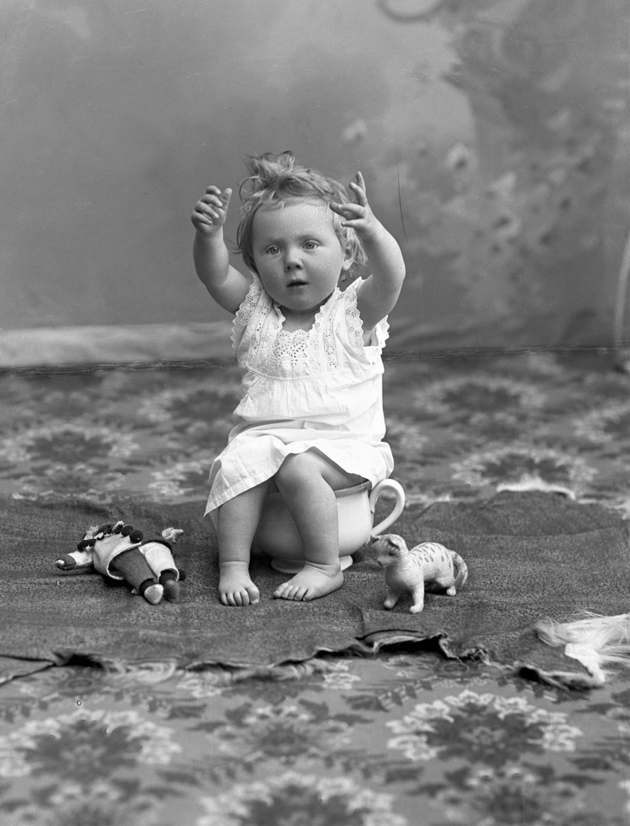 Atleljéporträtt av fotografens systerdotter, Anna-Stina Alling, som sitter på pottan och räcker upp händerna. På golvet ligger till vänster en (Pierrot)docka lllloch till höger en leksakskatt.