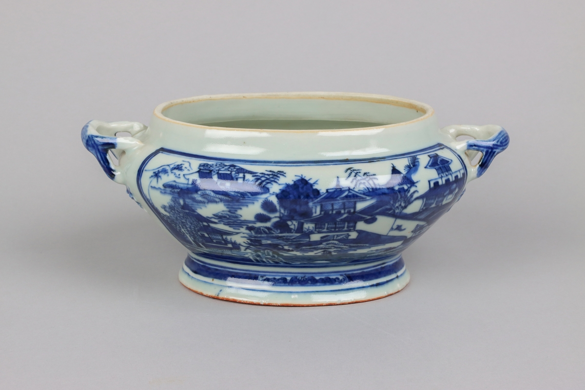 Oval, lobed bowl with angular handles, supported on a central foot. On the both sides decor of landscape scenes of pagodas, buildings, gardens and waters. Handles in shapes of bent tree branches. On the foot a  dark blue border with rectangular reserves filled with symbols of fortunes. All decor in blue underglaze. The base of the bowl  with unglazed ring.