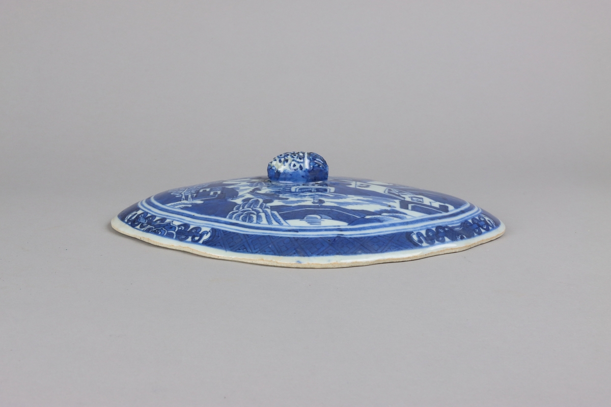 Oval form with a slightly domed form,  on top a knob in shape of a fruit. On the lid landscape scenes of pagodas, buildings,  bridges, figures with parasolls,  gardens and waters. The edge of the lid is decorated with a wide dark blue border with a criss cross pattern and reserves filled with symbols of good fortune. All decor in blue underglaze. The inside of the lid with out decoration.