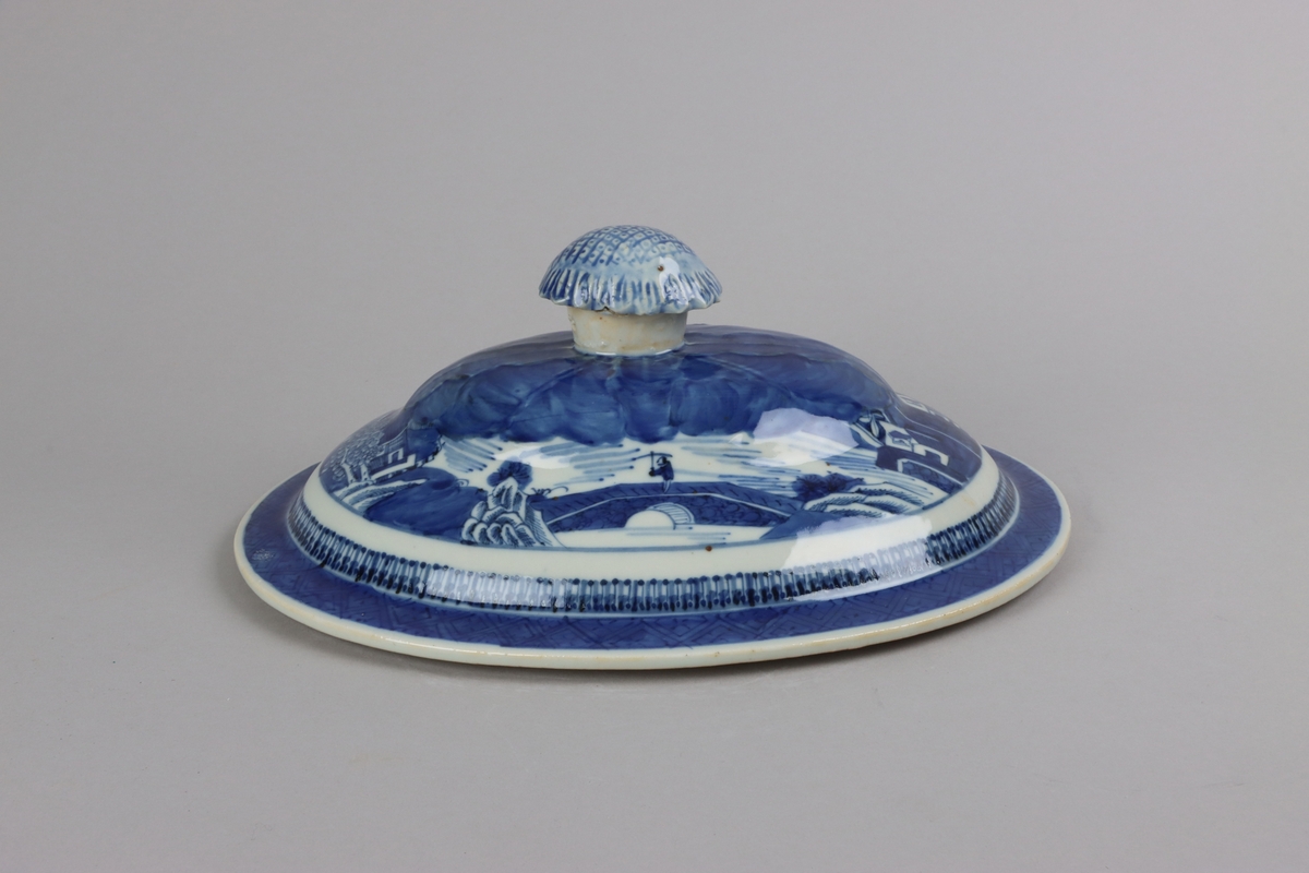 Oval shaped and slightly domed with knob in form of a flower head. On the top of lotus leaves and a list of pagoda lanscapes. The edge of the lid decorated with a dark blue  border in a criss cross pattern. All decor in blue underglaze. The inside of the lid is without decorations.