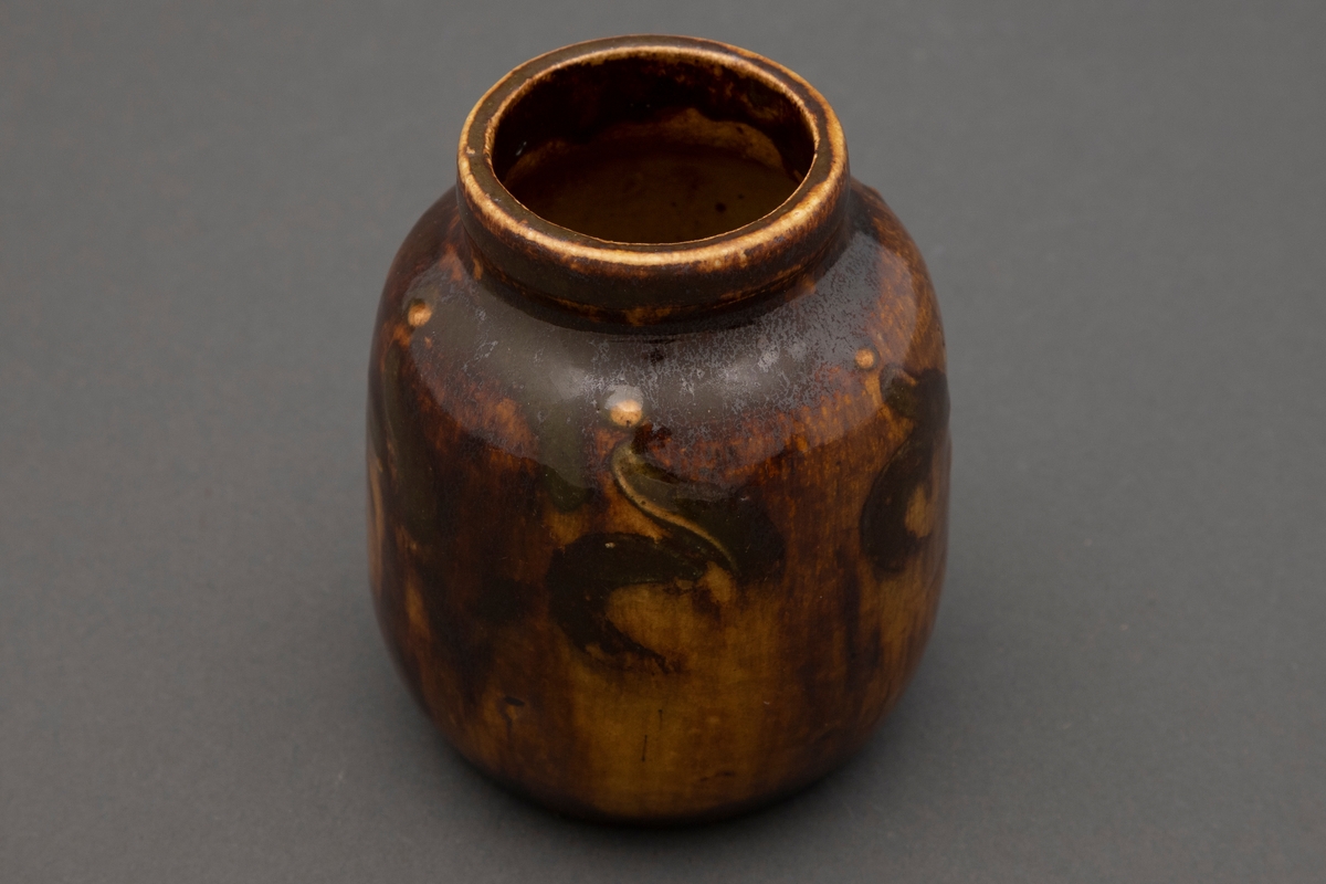Liten vase i leirgods. Vasen har en liten hals med markert utovergående kant. Gul med mørk brun flyteglasur. Dekorert med karakteristisk Art Nouveau-dekor.