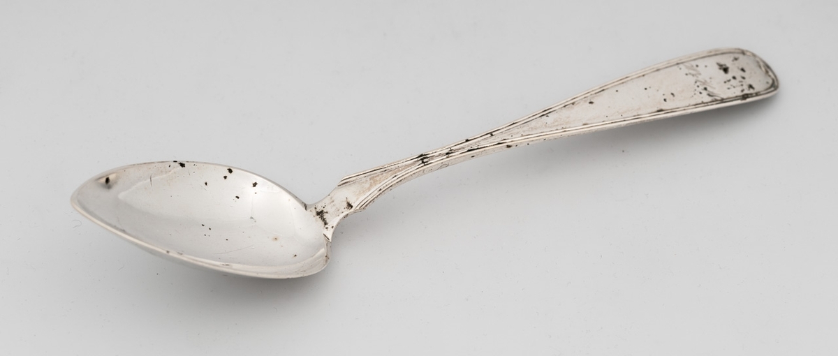 Skje i sølv med dråpeformet blad med spiss ende. Skaftet er buet med rille rundt kanten, samt avrundet avslutning.
